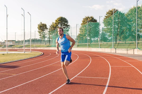 Young man, a professional athlete with a serious and focused face running along an outdoor athletic track near the sports field