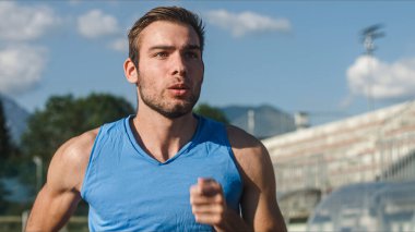 Genç bir adam, ciddi ve odaklanmış bir yüzü olan profesyonel bir atlet spor sahasının yakınındaki açık hava atletizm pistinde koşuyor.