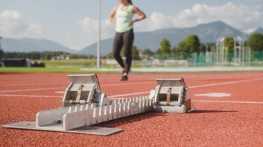 Başlangıç bloğu aygıtı atletik bir piste yerleştirilmiş, uzaktan bir kadın silueti yaklaşıyor, yakın çekim.