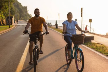 Caucasian couple enjoying a ride on beach cruiser bikes relaxed pedaling and talking near the beautiful sea illuminated by the afternoon sun