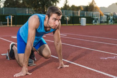 Mavi spor giyim giyen genç profesyonel erkek sporcu açık hava antrenmanı sırasında sprint koşusu için başlangıç pozisyonu alıyor.