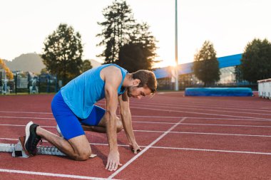 Mavi spor giyim giyen genç profesyonel erkek sporcu açık hava antrenmanı sırasında sprint koşusu için başlangıç pozisyonu alıyor.