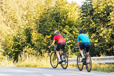 İki profesyonel bisikletçi, bir kadın ve bir adam, asfalt orman yolunda bir yarış sürüyorlar.