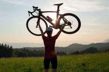 Gün batımında iki kolu başının üstünde bisiklet kaldıran kadın bisikletçi güzel yeşil doğayla birlikte. Kazanmak, başarı ve başarı kavramı.