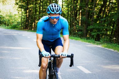 Beyaz erkek bisikletçi, profesyonel yarış ekipmanlarıyla açık yolda bisiklet sürerek ormandan geçiyor.