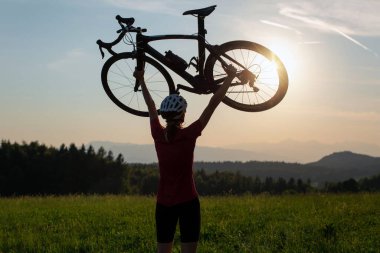 Gün batımında iki kolu başının üstünde bisiklet kaldıran kadın bisikletçi güzel yeşil doğayla birlikte. Kazanmak, başarı ve başarı kavramı.
