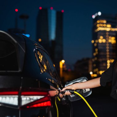 Gece bir iş merkezi gökdeleninde fazla mesai saatlerinde siyah elektrikli arabasını şarj eden genç bir kadın.