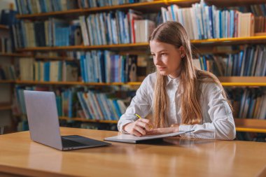 Akademik genç kız kütüphanede okuyor, ödevleri çözüyor, cevapları not defterine yazıyor ve siliyor. Bilgi ve okul kavramı.