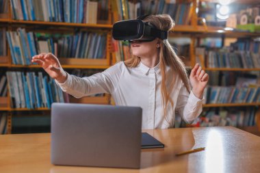 Kütüphanede modern teknoloji aletleri kullanan bir kız öğrenci, bir laptop ve VR kulaklık. Eğitimde sanal gerçeklik deneyimi kavramları.