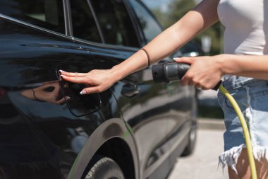 Mahalledeki bir elektrik istasyonunda zarif siyah EV arabasını şarj eden genç bir kadın. Temiz ve alternatif enerji kavramı.