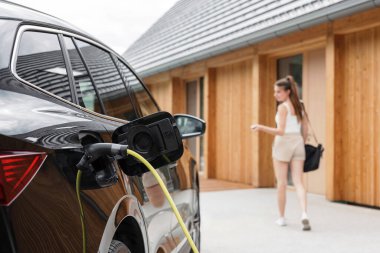 Gülümseyen kız siyah elektrikli bir arabanın şarj aletini fişe takıyor, ev kapısına giriyor ve uzaktan kumandayla aracı kilitliyor.