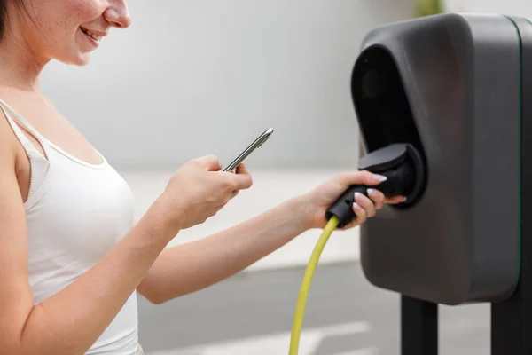 年轻女性 把充电器放在停放在停车场的电动汽车上 并在手机上调整电动汽车充电程序 — 图库照片