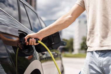 Adamın biri soketi açıyor ve şehir caddesindeki EV şarj istasyonunda siyah bir elektrikli arabaya şarj cihazı takıyor.