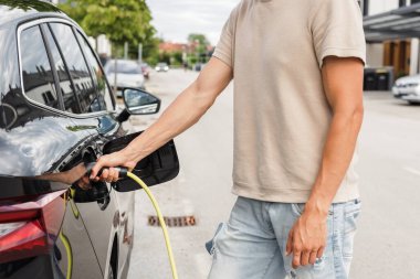 Yazlık kıyafetli bir adam, caddeye park etmiş elektrikli arabayı şarj ediyor. Yeşil ve sürdürülebilir enerji.