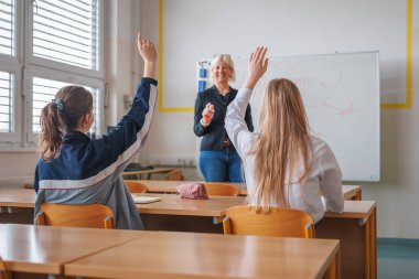 Kız öğrenci notlar yazar ve sınıftaki bir bayan öğretmen tahtaya otururken elini kaldırır. Sınıf ortamı kavramı.