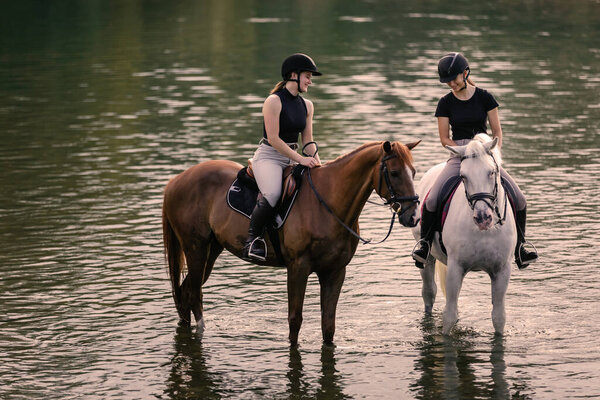 Riders, two young women riding beautiful horses down the calm river surrounded by the green grove