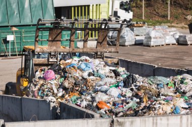 Yellow skid steer loader moving wooden waste material, shaking out a scrap grapple on the garbage heap in the materials recovery facility