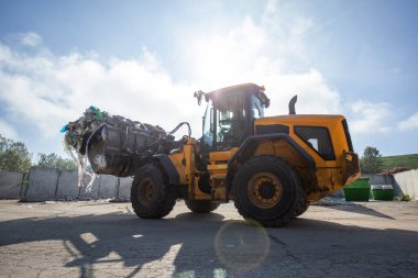 Yellow wheel loader, with lifted scrap grapple, moving along the recycling center area in process handling dumped waste