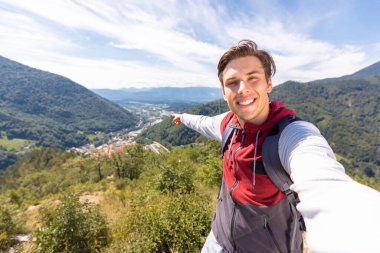 Gülümseyen adam bir selfie videosu çekiyor, konuşuyor ve doğal güzelliği işaret ediyor, arka planda dağ manzarası var.