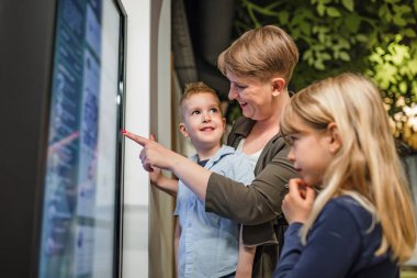 Aile, şirin sarışın çocuk ve küçük kız kardeşi interaktif dokunmatik ekran görsel yardım kullanarak anneleriyle müzede eğleniyorlar..