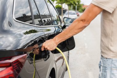 Yazlık kıyafetli bir adam, caddeye park etmiş elektrikli arabayı şarj ediyor. Yeşil ve sürdürülebilir enerji.