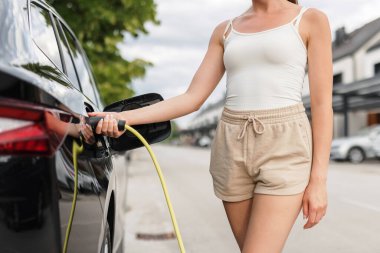 Günlük kıyafetli bir kız, caddeye park etmiş elektrikli arabayı şarj ediyor ve konut binasına gidiyor.