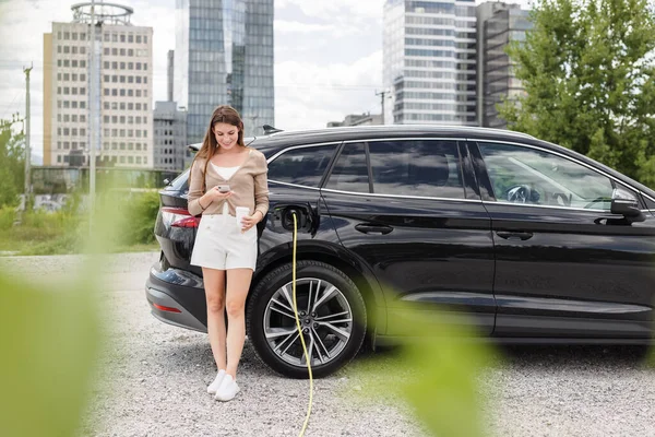 Woman charging electric car in city and adjusting EV charging app on smartphone