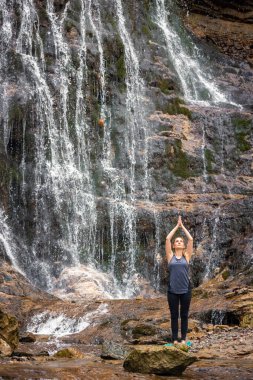 Sakin genç bir kadın güzel şelalede meditasyon yapıyor ve doğayla bağ kuruyor. Akış yogası kavramı.