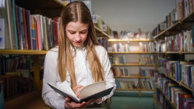 Okul kütüphanesindeki kitaplıkların arasına girip okumak için ilham verici bir kitap seçen neşeli, neşeli, sevimli okul kızı.