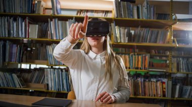 Okul kütüphanesinde ders çalışırken laptop ve sanal gerçeklik gözlüğü kullanan genç bir kız. Yeni eğitim teknolojisi kavramı.