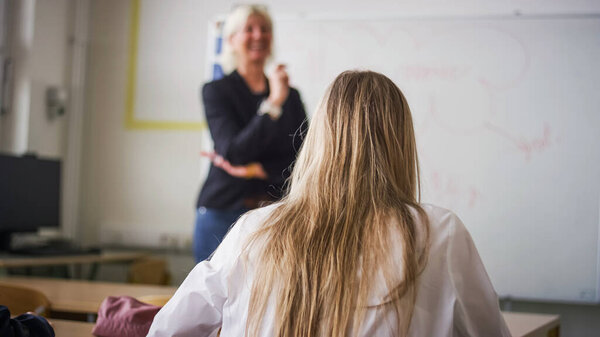 Девочка-подросток сидит за школьным столом в классе и слушает учителя