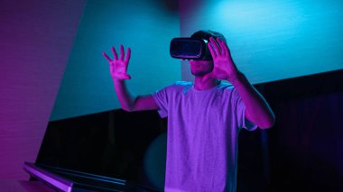 VR kulaklıklı genç adam mor ışıklı bir odada sanal gerçeklik deneyimi yaşıyor, havada çeşitli el hareketleri yapıyor..