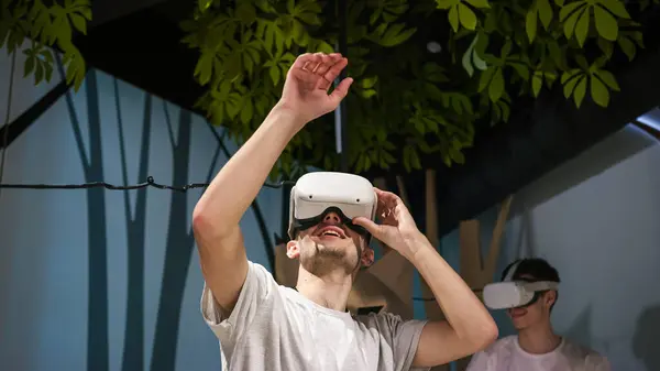 Interactive experience in the virtual reality room, two male students using VR glasses, looking around the space and moving arms through the air.