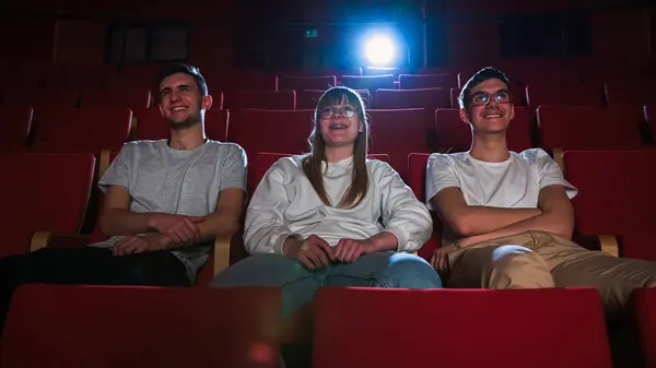 Three friends in the cinema hall, a girl and two boys having fun watching a film in the empty movie place. Entertainment and people concept.
