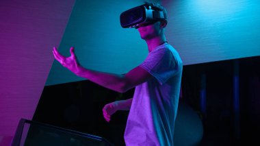 VR kulaklıklı genç adam mor ışıklı bir odada sanal gerçeklik deneyimi yaşıyor, havada çeşitli el hareketleri yapıyor..