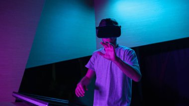 VR kulaklıklı genç adam mor ışıklı bir odada sanal gerçeklik deneyimi yaşıyor, havada çeşitli el hareketleri yapıyor..