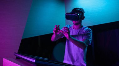 VR kulaklıklı genç adam mor ışıklı bir odada sanal gerçeklik deneyimi yaşıyor, havada çeşitli el hareketleri yapıyor..