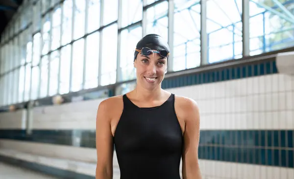 Portrait of a woman, a professional swimmer in black one-piece swimwear and a black cap, looking at the camera and putting on swimming goggles.