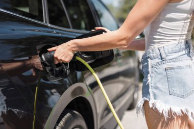 Şehir merkezinden gülümseyen kız geliyor ve elektrikli arabasının şarj kablosunun fişini çekiyor. Kentsel yaşam ve modern yaşam tarzı kavramı.