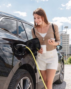 Şehir iş merkezinin yakınındaki otoparkta elektrikli arabasını şarj eden kız arkada gökdelenler görünüyor.