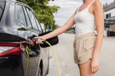 Mahalledeki bir elektrik istasyonunda zarif siyah EV arabasını şarj eden genç bir kadın. Temiz ve alternatif enerji kavramı.