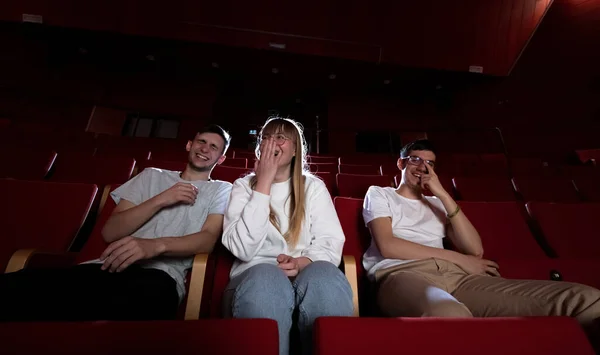 Friends, a girl, and two boys enjoying a comedy movie in the cinema, having fun and happily laughing during film projection.