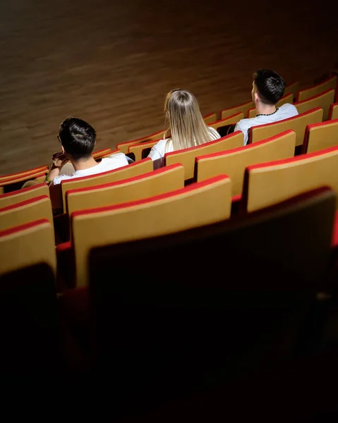 Three friends in the cinema hall, a girl and two boys having fun watching a film in the empty movie place. Entertainment and people concept.