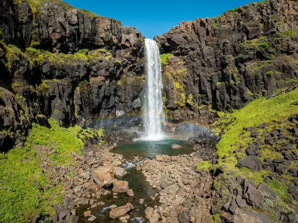 Stunning Iceland waterfall with icy cold water and the pool at the base filled with rocks on a sunny day, aerial view. Travel destination and nature wonder concepts.