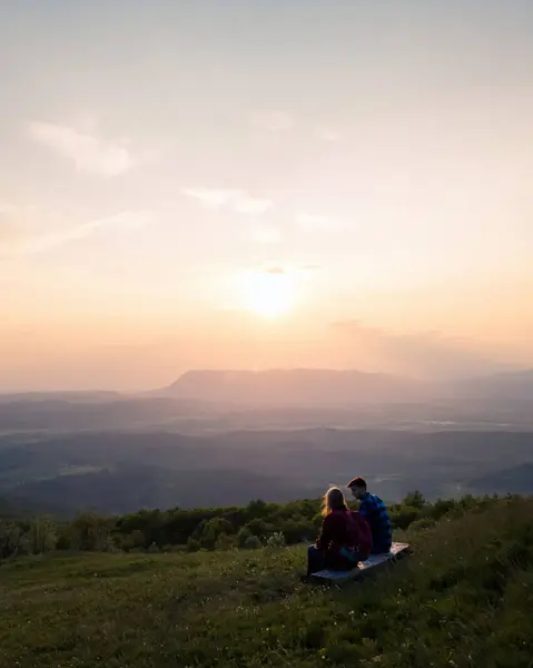 Young couple enjoying the fantastic mountain panorama at sunset while sitting on the bench at a viewpoint. Picturesque moments and natural beauty concepts.