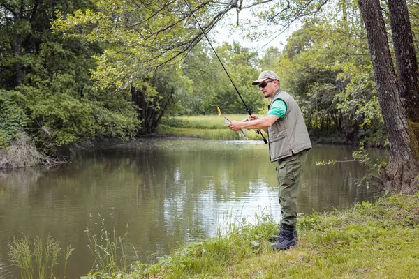 An angler standing on a riverbank and successfully catching a fish during freshwater fishing season. Concepts of individual sport and leisure activity.
