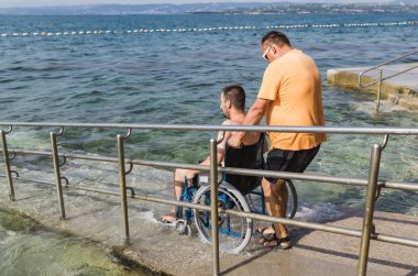 Erkek asistan, tekerlekli sandalyedeki bir adama denize açılan bir rampada denizin tadını çıkarması için yardım ediyor. Erişilebilir plaj kavramı.