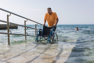 Erkek asistan, tekerlekli sandalyedeki bir adama denize açılan bir rampada denizin tadını çıkarması için yardım ediyor. Erişilebilir plaj kavramı.
