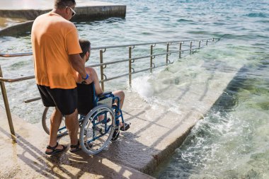 Tekerlekli sandalye kullanıcısı erişilebilir plaj rampasında bir asistanın yardımıyla denize giriyor. Engellilik ve turizm kavramları.