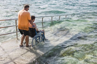 Tekerlekli sandalye kullanıcısı erişilebilir plaj rampasında bir asistanın yardımıyla denize giriyor. Engellilik ve turizm kavramları.